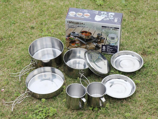 Eight pieces of stainless steel camping cookware are neatly arranged on a white surface. The set includes two pots (one larger than the other) with foldable handles extended, a frying pan with a foldable handle, a lid, two plates, and two cups with handles.