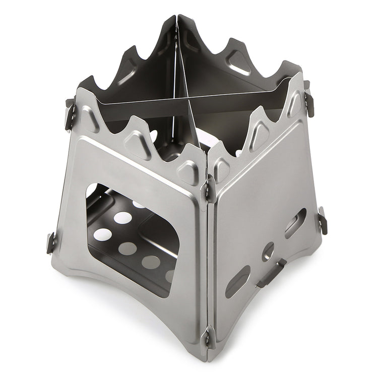 A small, silver stainless steel wood-burning stove is shown unfolded against a white background. The stove has a boxy structure with air vents and a cross-shaped trivet on top.