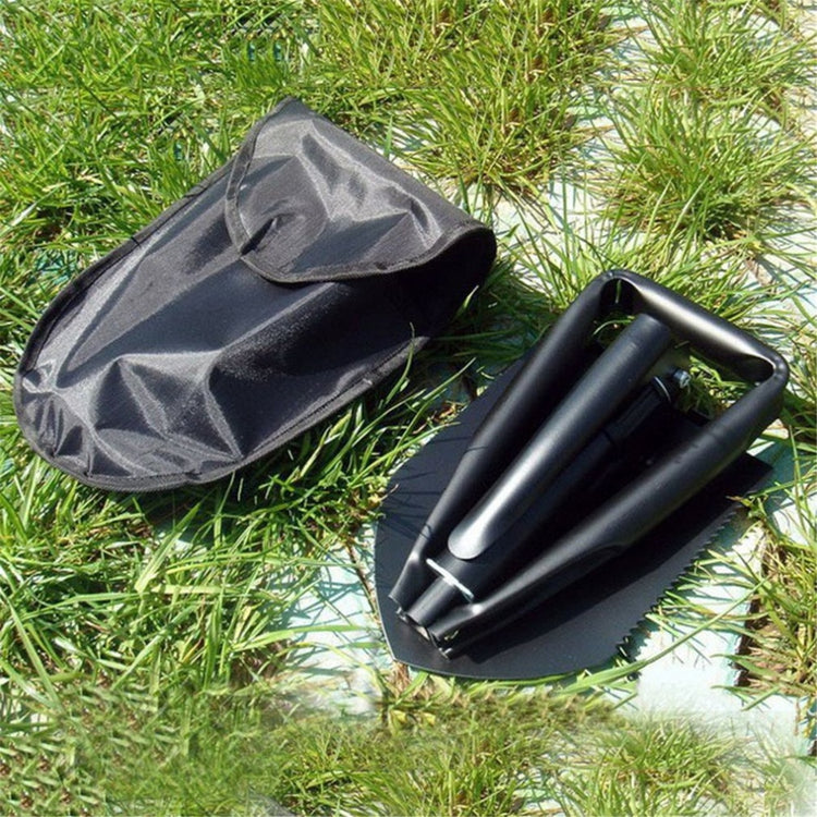 A black carbon steel folding shovel is shown folded next to its black nylon carry pouch on artificial green grass. The shovel has a triangular blade and a D-shaped handle.