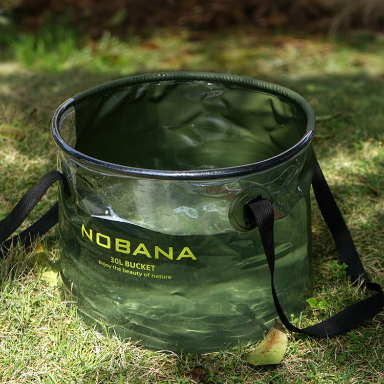 A translucent army green Nobana 30L folding bucket filled with water sits on green grass. The bucket has black trim and black carrying straps. The Nobana logo and "30L BUCKET Enjoy the beauty of nature" are printed in yellow on the side.