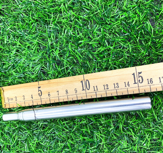 A silver expandable stainless steel fire tube is shown extended and lying on artificial green grass next to a wooden ruler measuring in centimeters. The fire tube is approximately 15 cm when collapsed.