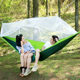 A two-tone (light green and dark green) ultralight parachute hammock with a white mosquito net suspended above it. Below are two silver carabiners, two black tree straps, a bundle of white rope, and a green drawstring storage bag.