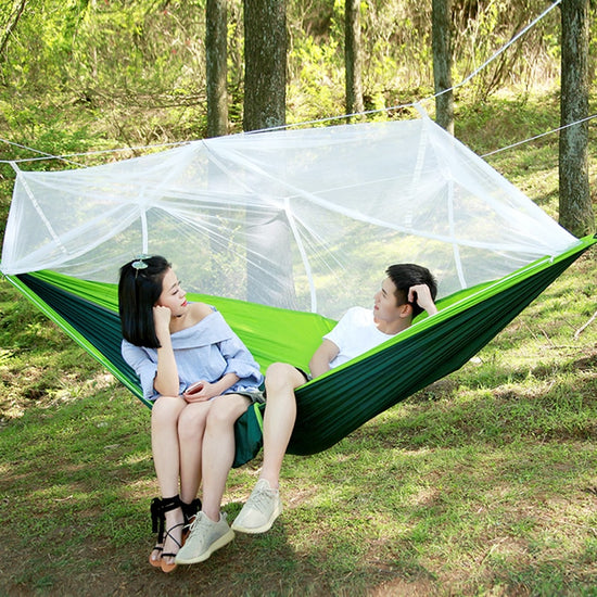 A two-tone (light green and dark green) ultralight parachute hammock with a white mosquito net suspended above it. Below are two silver carabiners, two black tree straps, a bundle of white rope, and a green drawstring storage bag.