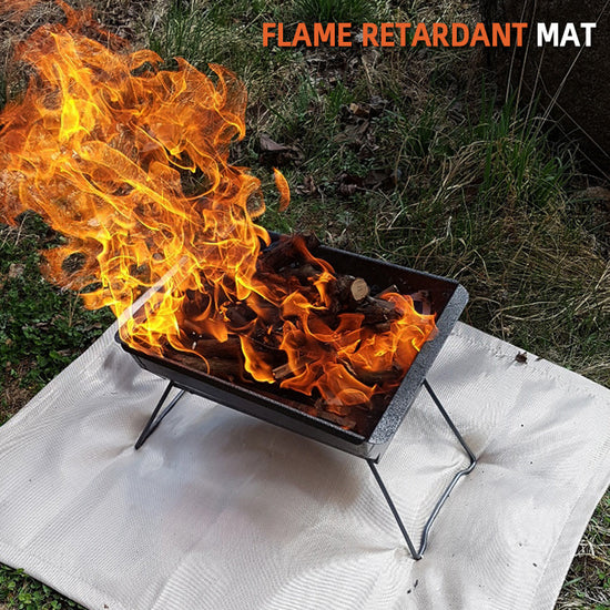 A portable fire pit with bright flames burning on top of a white flame-retardant mat placed on grassy ground, illustrating its use for protecting the ground from heat damage during outdoor camping.