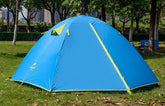 A blue Naturehike 2-person double-layer camping tent is pitched on green grass with trees in the blurred background. The tent has yellow accents around the zippers and ventilation flaps, and the Naturehike logo is visible on the side.