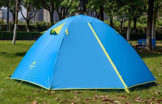 A blue Naturehike 2-person double-layer camping tent is pitched on green grass with trees in the blurred background. The tent has yellow accents around the zippers and ventilation flaps, and the Naturehike logo is visible on the side.