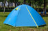 A blue Naturehike 2-person double-layer camping tent is pitched on green grass with trees in the blurred background. The tent has yellow accents around the zippers and ventilation flaps, and the Naturehike logo is visible on the side.