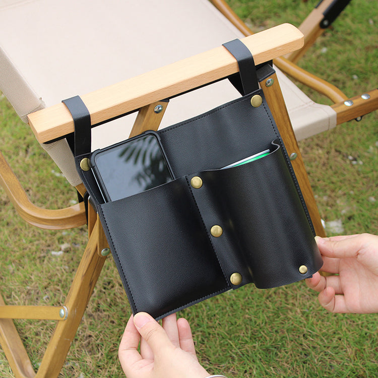 A green leather cylindrical storage bag is shown attached to the armrest of a chair. A black smartphone is visible in one pocket, and a green and silver beverage can is in the other cylindrical holder. The background is a white marble-like surface with a framed picture and books slightly out of focus.
