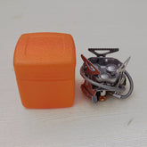  A compact silver camping gas stove with three burner heads and foldable pot supports is shown next to its bright orange plastic carrying case on a light wooden surface.