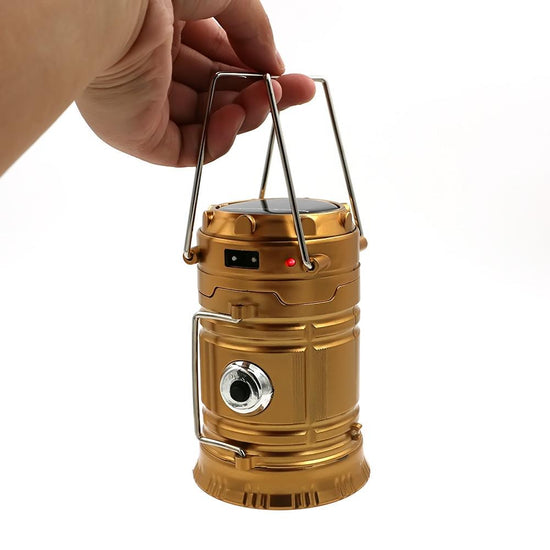 A gold-colored collapsible LED lantern is shown being held by a hand against a white background. The lantern is partially extended, showing its cylindrical shape with a carrying handle at the top and a lens visible on the side. The top of the lantern has a solar panel.