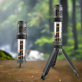 Two cylindrical LED camping lights are shown against a blurred natural background of trees and possibly a waterfall. The light on the left stands upright on its own. The light on the right is identical but is mounted on a small, black tripod with its legs extended. Both lights have a dark grey top section, a clear cylindrical middle section revealing orange internal components, and a dark grey base.