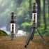 Two cylindrical LED camping lights are shown against a blurred natural background of trees and possibly a waterfall. The light on the left stands upright on its own. The light on the right is identical but is mounted on a small, black tripod with its legs extended. Both lights have a dark grey top section, a clear cylindrical middle section revealing orange internal components, and a dark grey base.