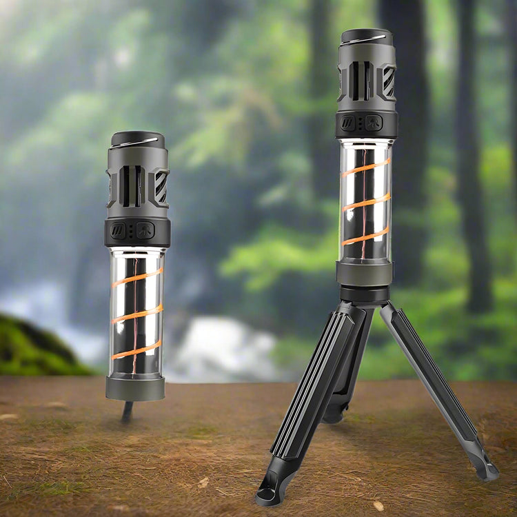 Two cylindrical LED camping lights are shown against a blurred natural background of trees and possibly a waterfall. The light on the left stands upright on its own. The light on the right is identical but is mounted on a small, black tripod with its legs extended. Both lights have a dark grey top section, a clear cylindrical middle section revealing orange internal components, and a dark grey base.