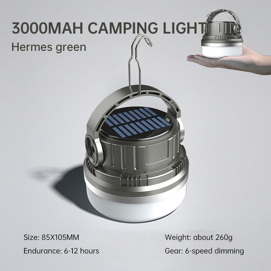 A round, silver and white camping lantern with a black solar panel on top and a silver hanging hook extended. Text above the lantern reads "3000MAH CAMPING LIGHT" and "Hermes green". Smaller text below indicates size, endurance, weight, and dimming modes. A hand holding a smaller version of the same lantern is visible in the upper right corner.