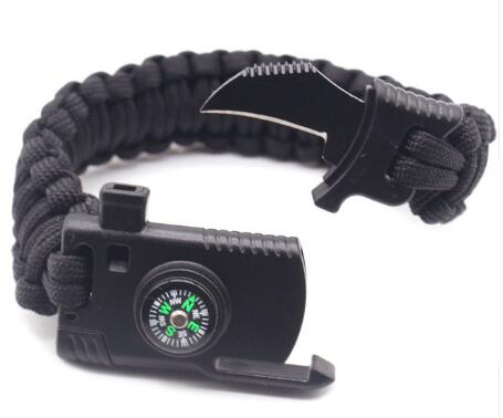 A black braided paracord survival bracelet is shown partially unbuckled, revealing a small knife blade, a compass on the buckle, and the ridged edge of a fire starter.