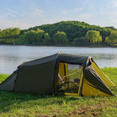A large, army green tunnel tent is pitched on grassy ground near a body of water and trees under a bright sky. The tent has a spacious main section with an open entrance revealing chairs inside, and a smaller, angled section extending from the front.