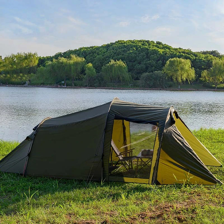 A large, army green tunnel tent is pitched on grassy ground near a body of water and trees under a bright sky. The tent has a spacious main section with an open entrance revealing chairs inside, and a smaller, angled section extending from the front.