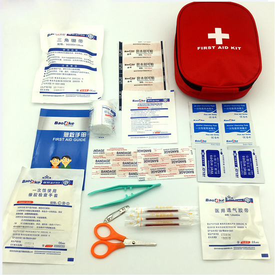 A red zippered first aid kit bag with a white cross on the front is shown open, revealing various medical supplies including bandages, antiseptic wipes, gauze pads, tape, scissors, tweezers, safety pins, and a first aid guide, all arranged on a white surface.