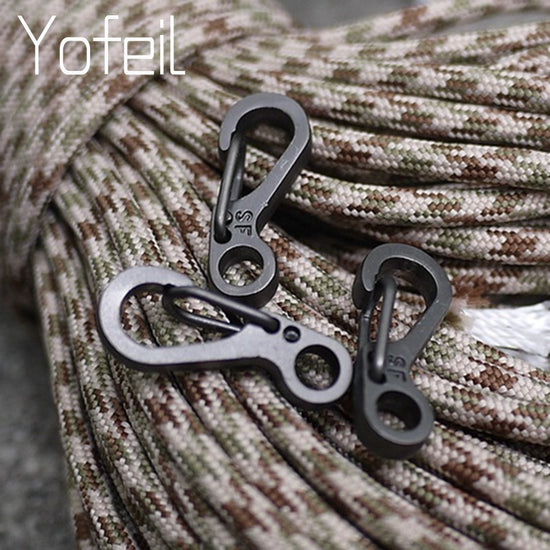 A close-up of three small, gun-gray aluminum alloy SF spring clip carabiners resting on a background of tan and brown camouflage paracord.