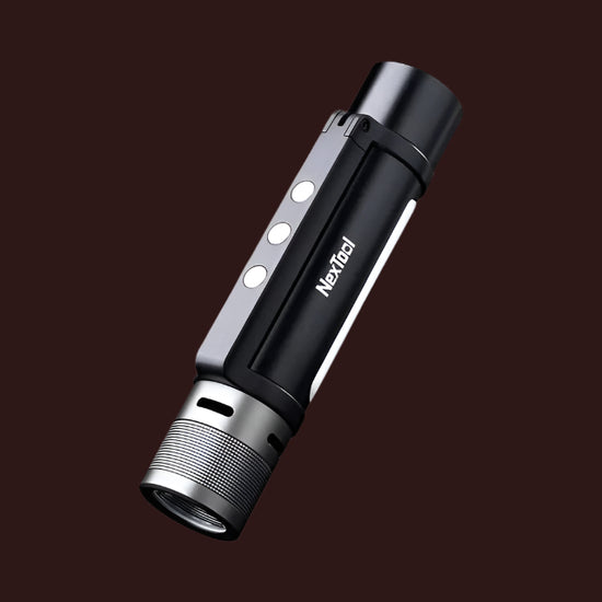 A black and silver NEXTOOL 6-in-1 flashlight is shown diagonally against a dark brown background. The flashlight has a textured silver head, a black body with the "NexTool" logo printed in white, three white indicator lights, and a lanyard attachment point at the base.