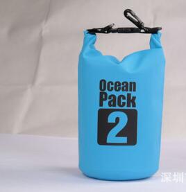 A light blue "Ocean Pack 2" branded waterproof dry bag is shown standing upright. The top of the bag is rolled and clipped shut with a black buckle. The background is a blurry light grey.