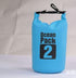 A light blue "Ocean Pack 2" branded waterproof dry bag is shown standing upright. The top of the bag is rolled and clipped shut with a black buckle. The background is a blurry light grey.