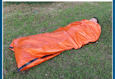 A person wrapped in a bright orange thermal emergency sleeping bag lies on green grass.