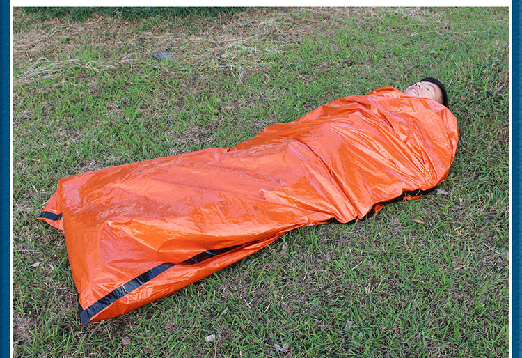 A person wrapped in a bright orange thermal emergency sleeping bag lies on green grass.