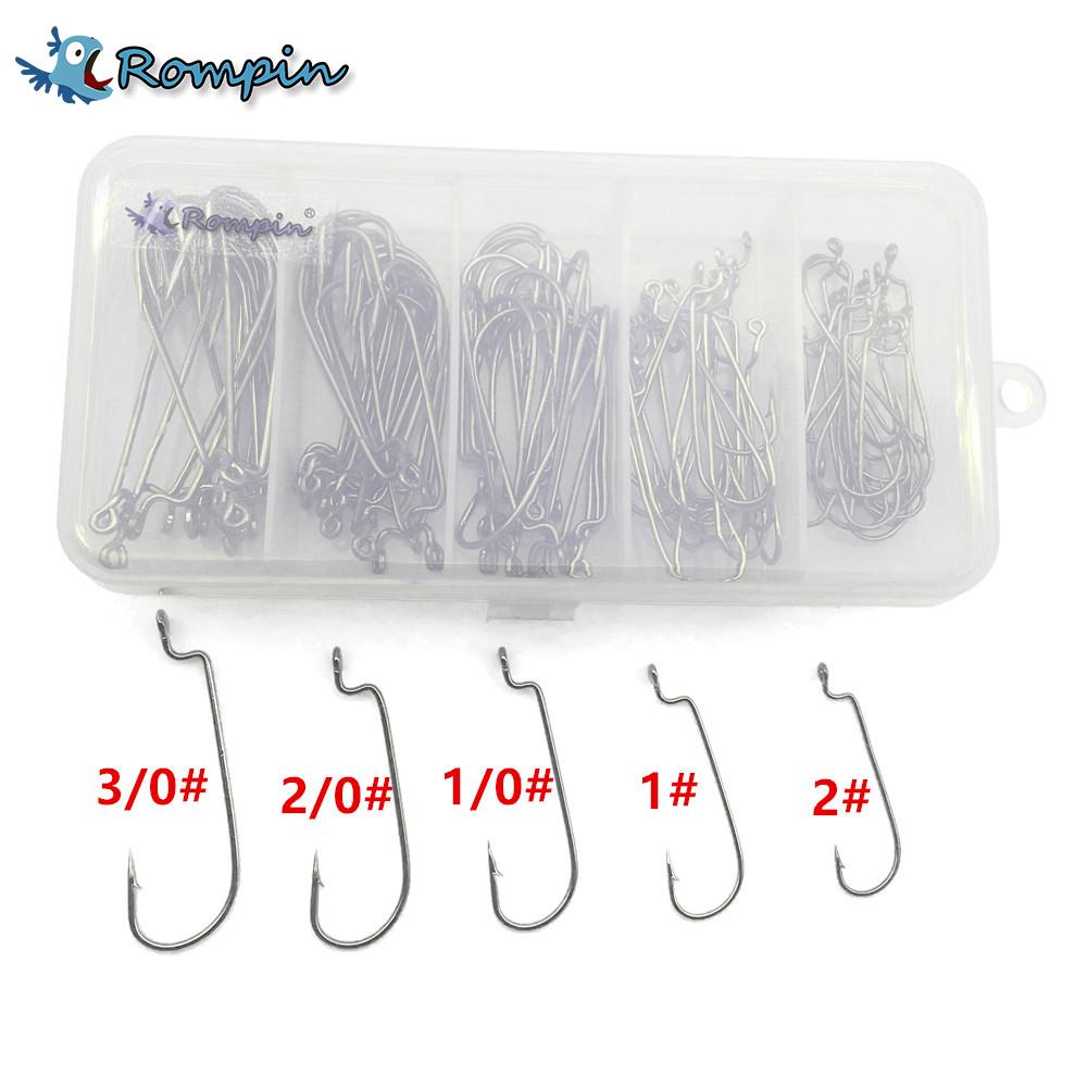 A clear plastic box containing approximately 100 silver jig head fishing hooks of various sizes, organized into five compartments. Below the box are individual hooks labeled with sizes 3/0