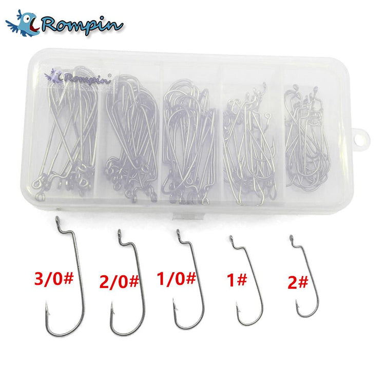 A clear plastic box containing approximately 100 silver jig head fishing hooks of various sizes, organized into five compartments. Below the box are individual hooks labeled with sizes 3/0